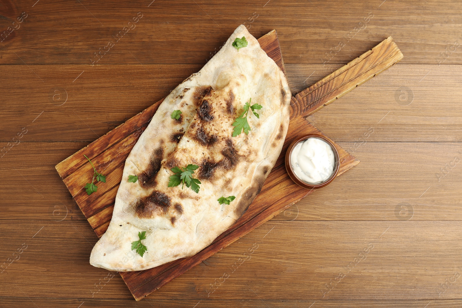 Photo of Board with tasty calzone, parsley and sauce on wooden table, top view