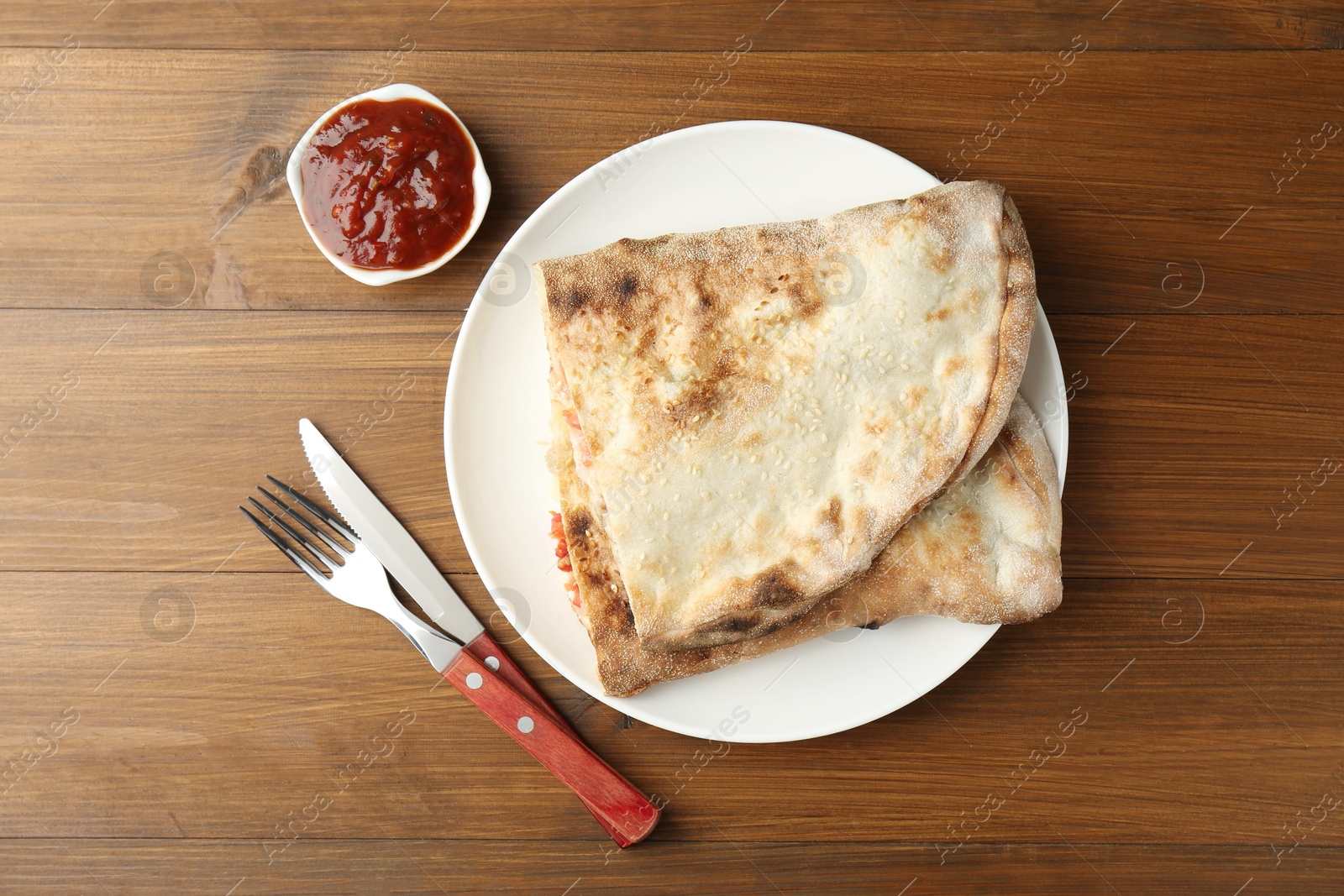 Photo of Halves of tasty calzone served with sauce on wooden table, flat lay