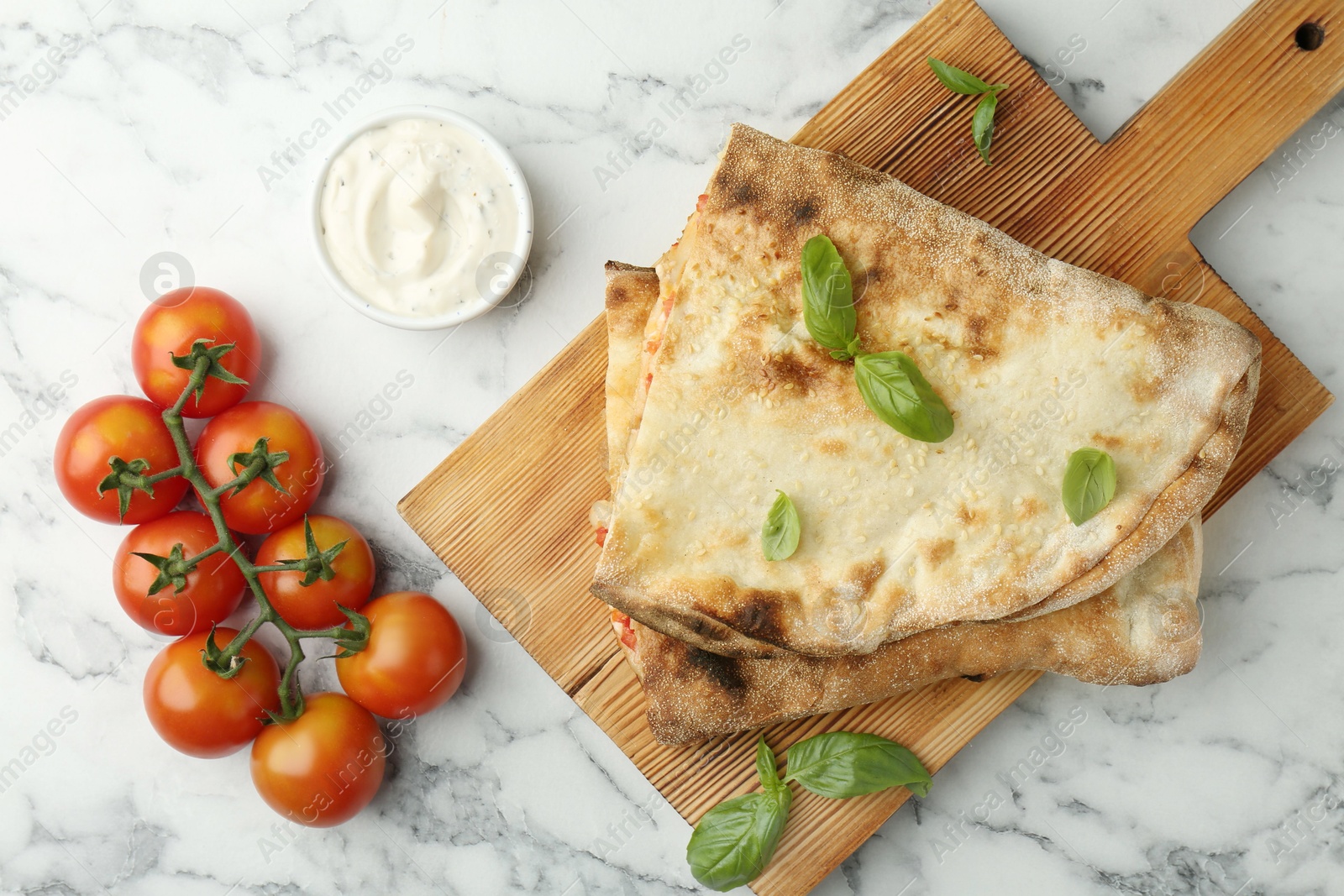 Photo of Halves of tasty calzone, basil, sauce and tomato on white marble table, flat lay