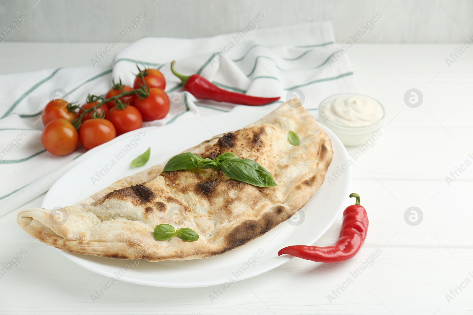 Photo of Tasty calzone, basil, chili pepper, sauce and tomatoes on white wooden table, closeup