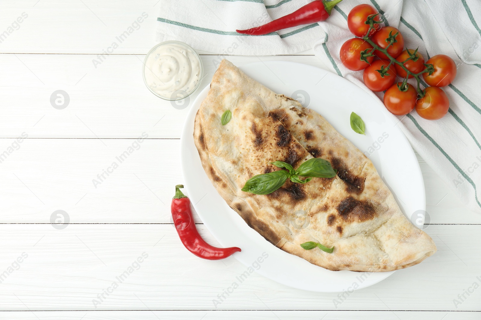 Photo of Tasty calzone, basil, chili pepper, sauce and tomatoes on white wooden table, flat lay. Space for text