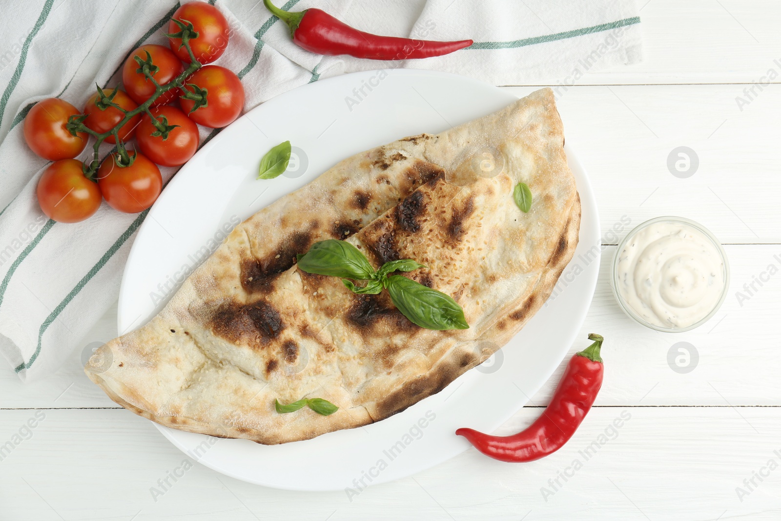 Photo of Tasty calzone, basil, chili pepper, sauce and tomatoes on white wooden table, flat lay