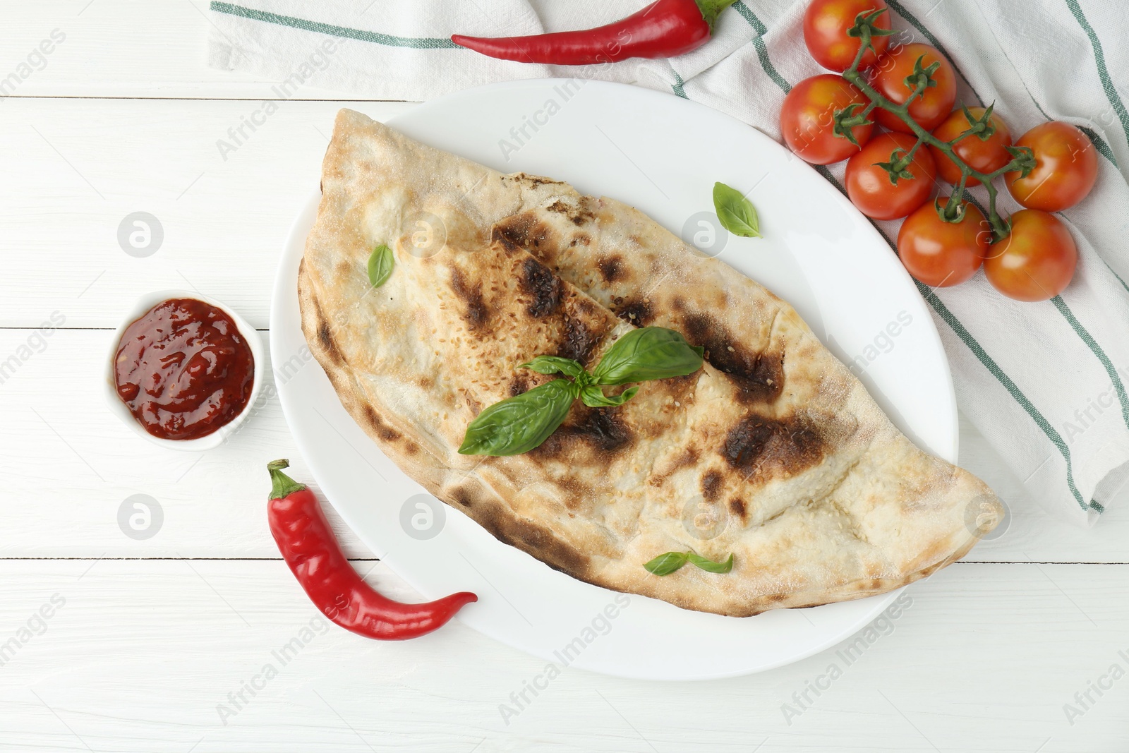 Photo of Tasty calzone, basil, chili pepper, sauce and tomatoes on white wooden table, flat lay