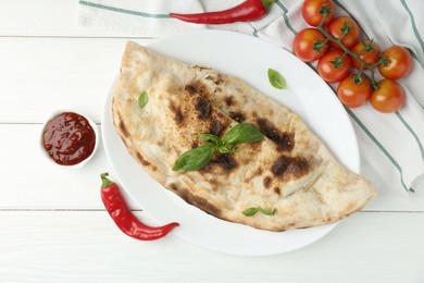 Photo of Tasty calzone, basil, chili pepper, sauce and tomatoes on white wooden table, flat lay