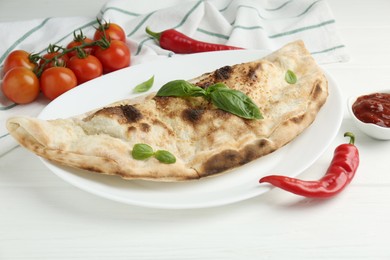 Photo of Tasty calzone, basil, chili pepper, sauce and tomatoes on white wooden table, closeup