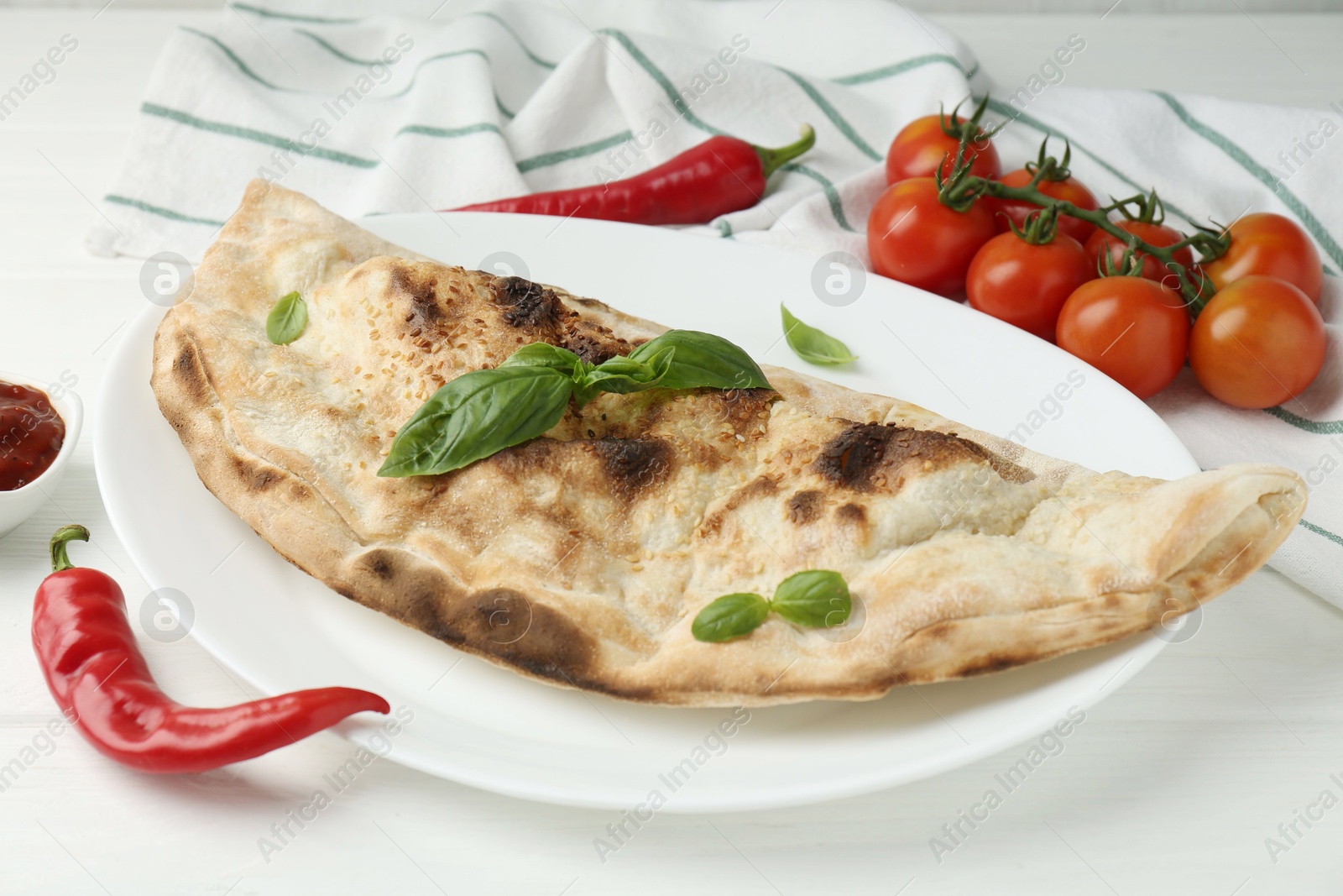 Photo of Tasty calzone, basil, chili pepper, sauce and tomatoes on white wooden table, closeup