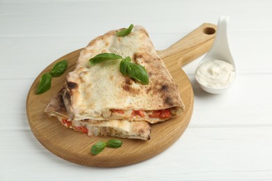 Photo of Halves of tasty calzone with meat, cheese, tomato, basil and sauce on white wooden table, closeup