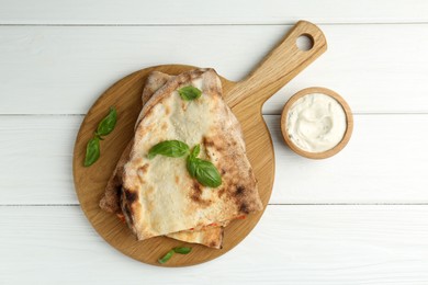 Photo of Halves of tasty calzone, basil and sauce on white wooden table, flat lay