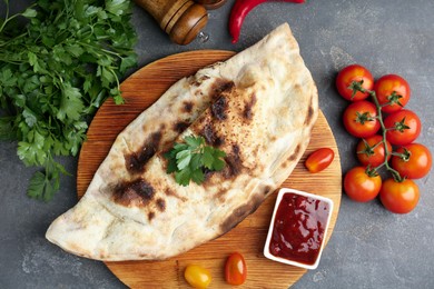 Photo of Board with tasty calzone, tomatoes, sauce and parsley on grey textured table, flat lay