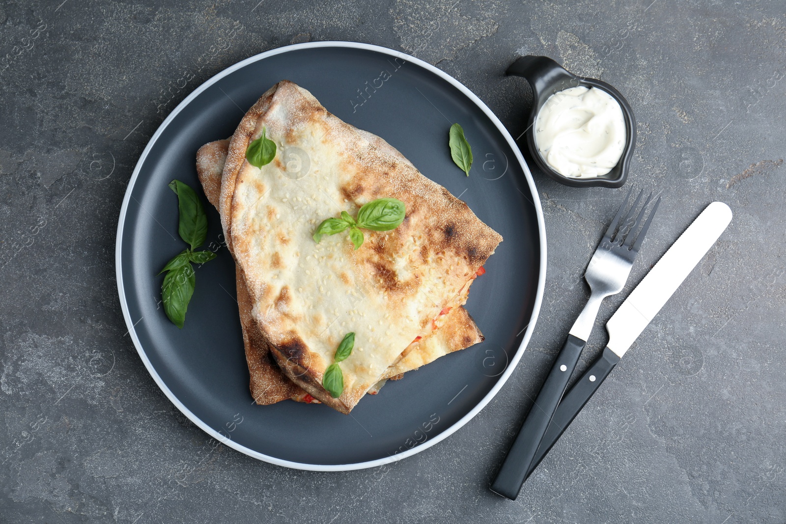 Photo of Halves of tasty calzone with basil served with sauce on grey textured table, flat lay