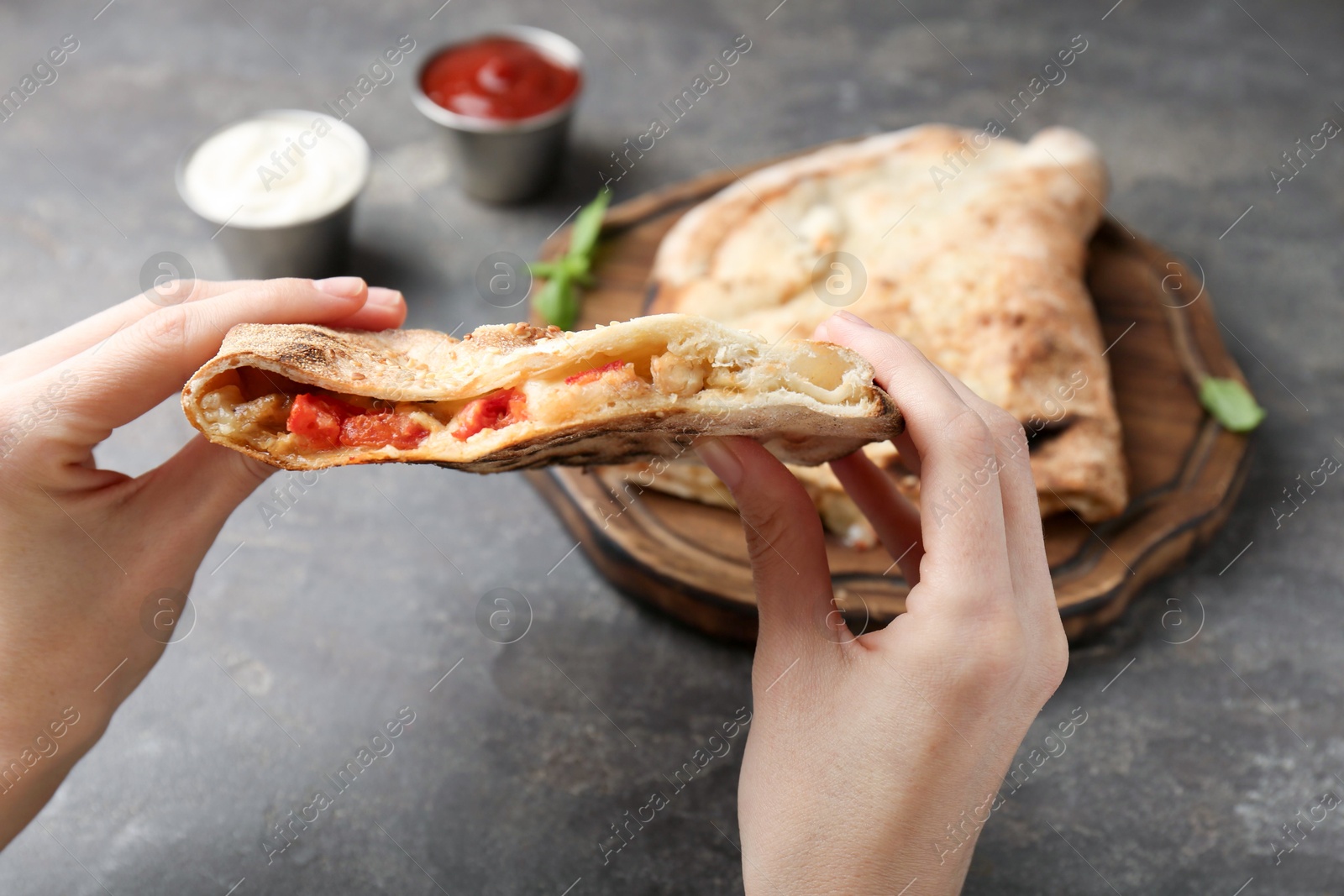 Photo of Woman eating tasty calzone with meat, cheese and tomato at grey textured table, closeup