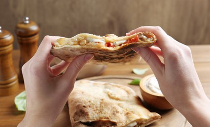 Photo of Woman eating tasty calzone with meat, cheese and tomato at wooden table, closeup