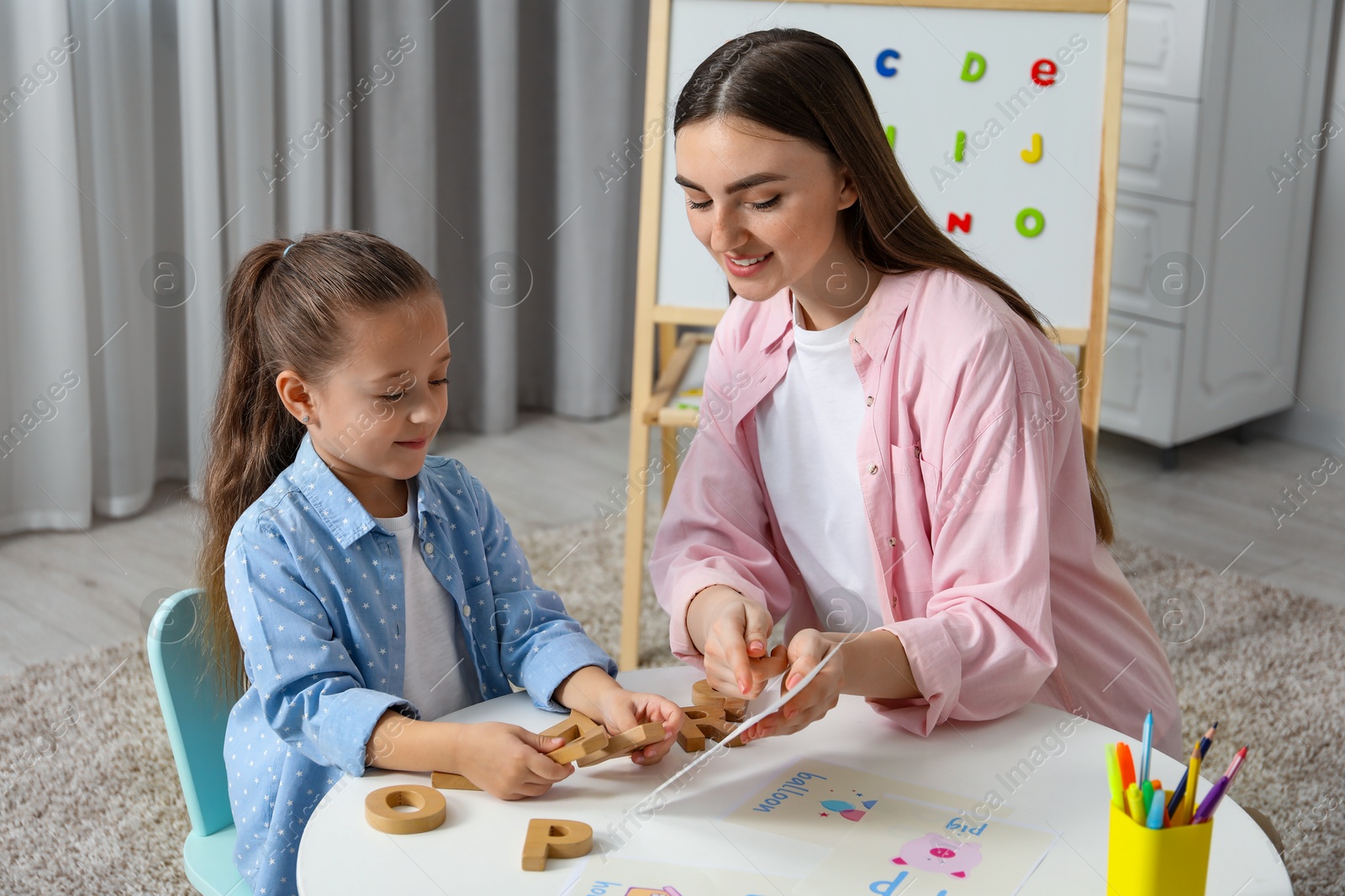 Photo of Speech therapist teaching little girl alphabet at white table indoors