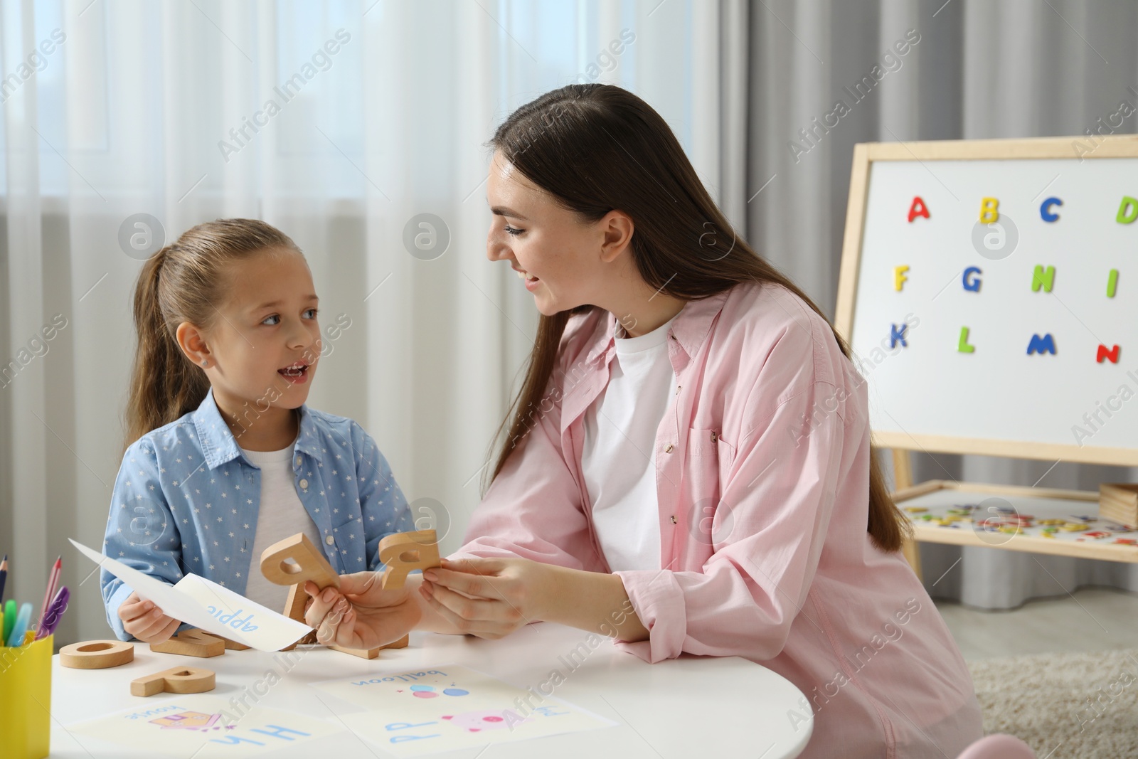 Photo of Speech therapist teaching little girl alphabet with wooden letters at white table indoors