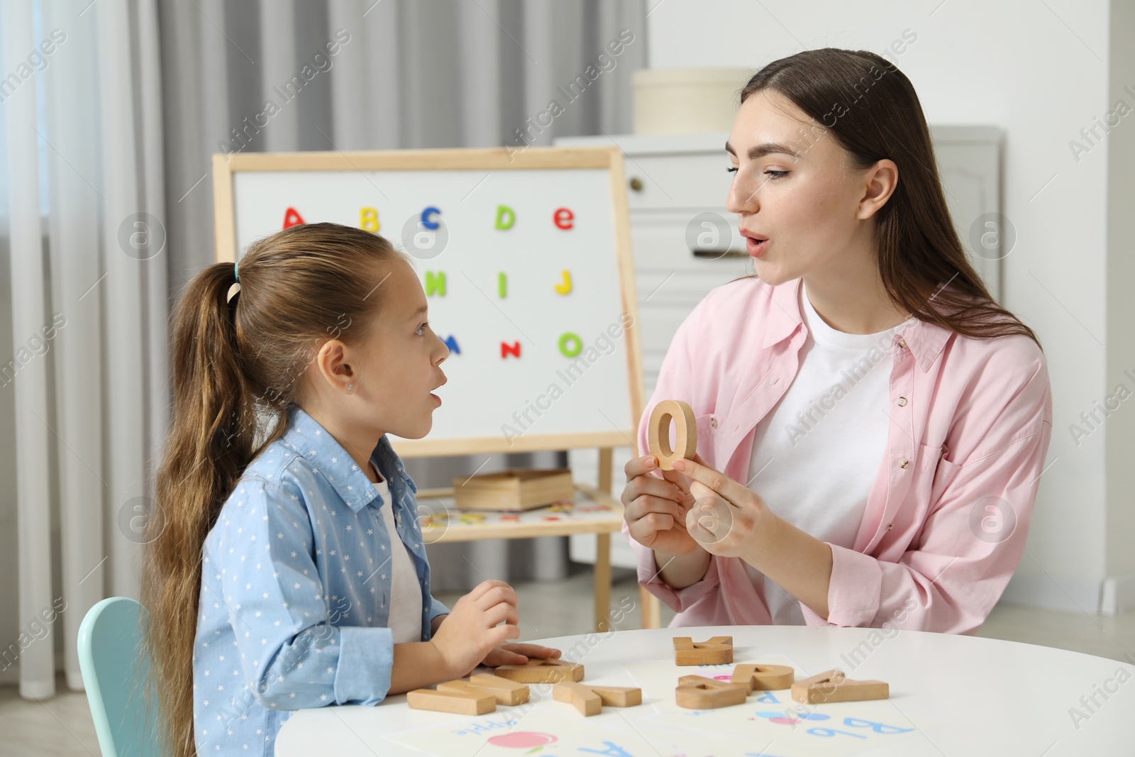 Photo of Speech therapist teaching little girl alphabet with wooden letters at white table indoors