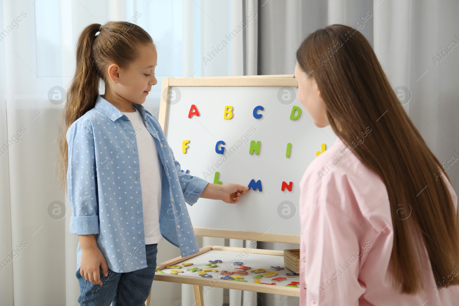 Photo of Speech therapist teaching little girl alphabet with magnetic letters indoors