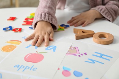 Photo of Little girl learning alphabet with different letters and pictures at white table indoors, closeup