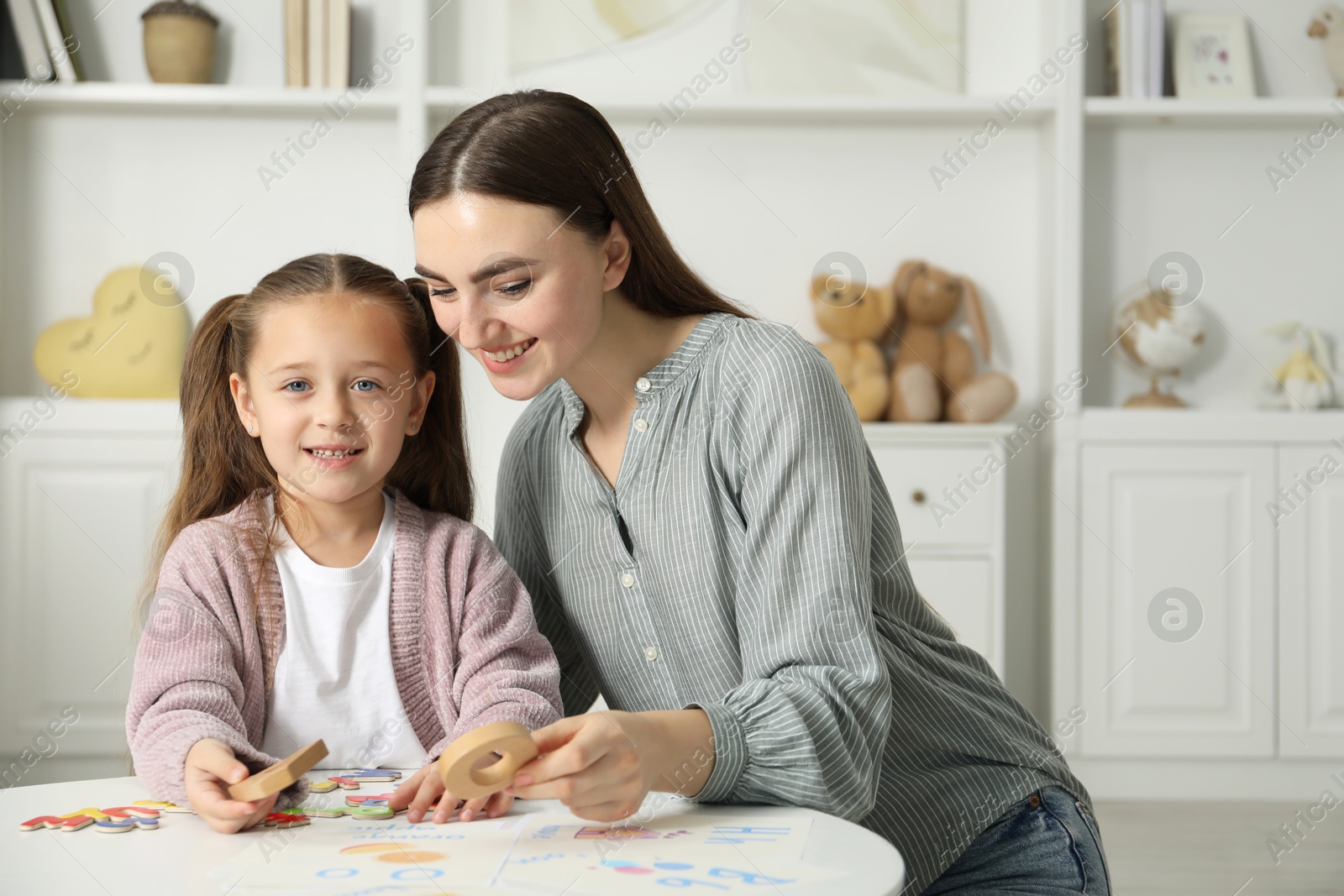 Photo of Speech therapist teaching little girl alphabet with different letters at white table indoors