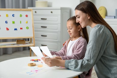 Photo of Speech therapist teaching little girl alphabet at white table indoors