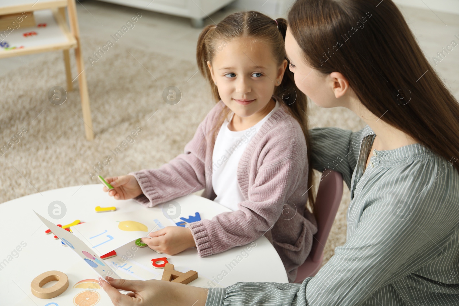 Photo of Speech therapist teaching little girl alphabet at white table indoors