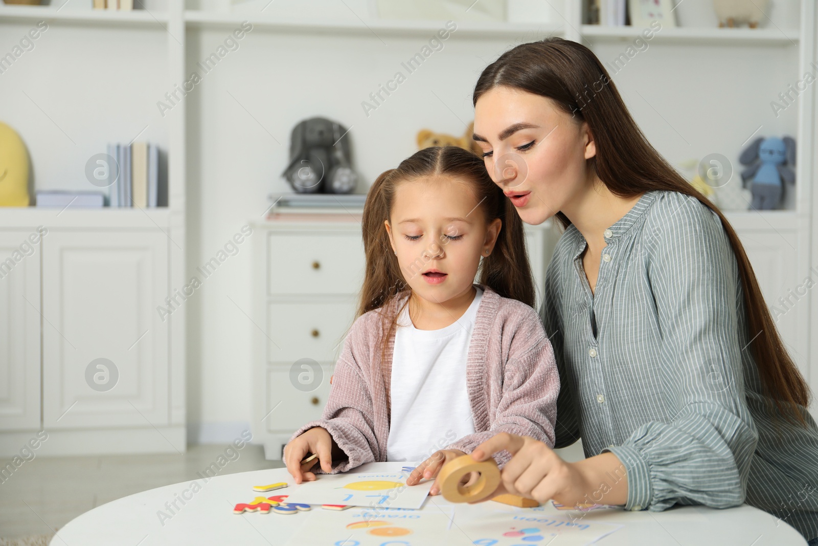 Photo of Speech therapist teaching little girl alphabet at white table indoors