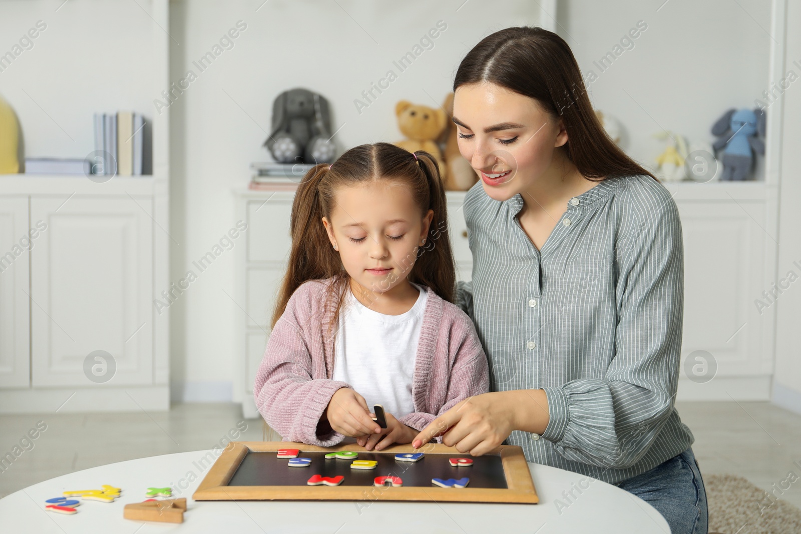 Photo of Speech therapist teaching little girl alphabet with magnetic letters at white table indoors