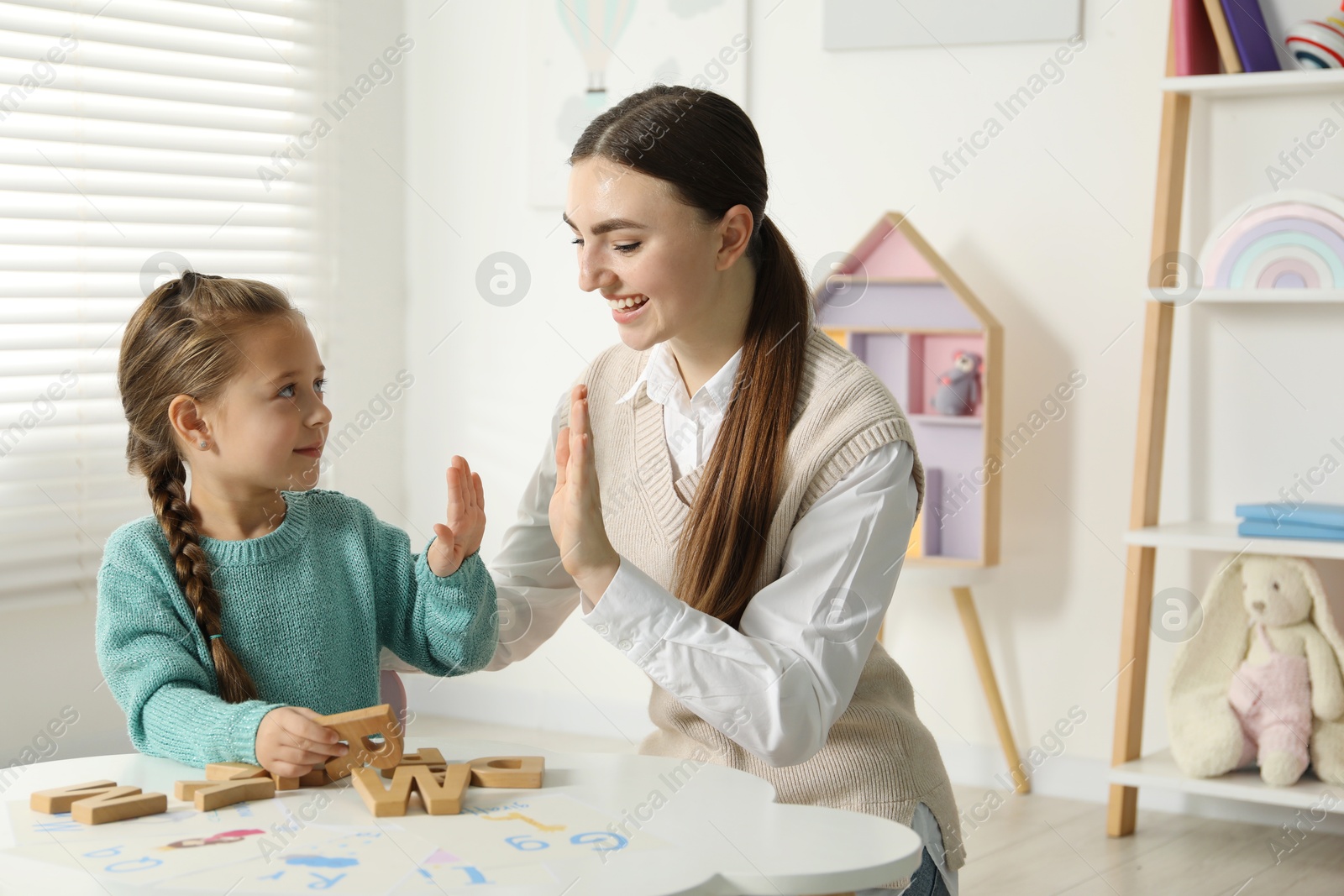 Photo of Speech therapist teaching little girl alphabet at white table indoors