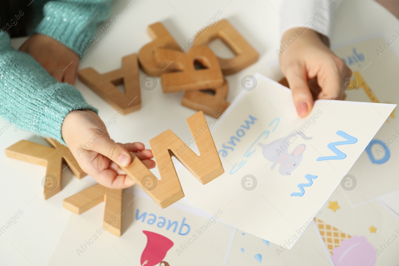 Photo of Speech therapist teaching little girl alphabet at white table indoors, closeup