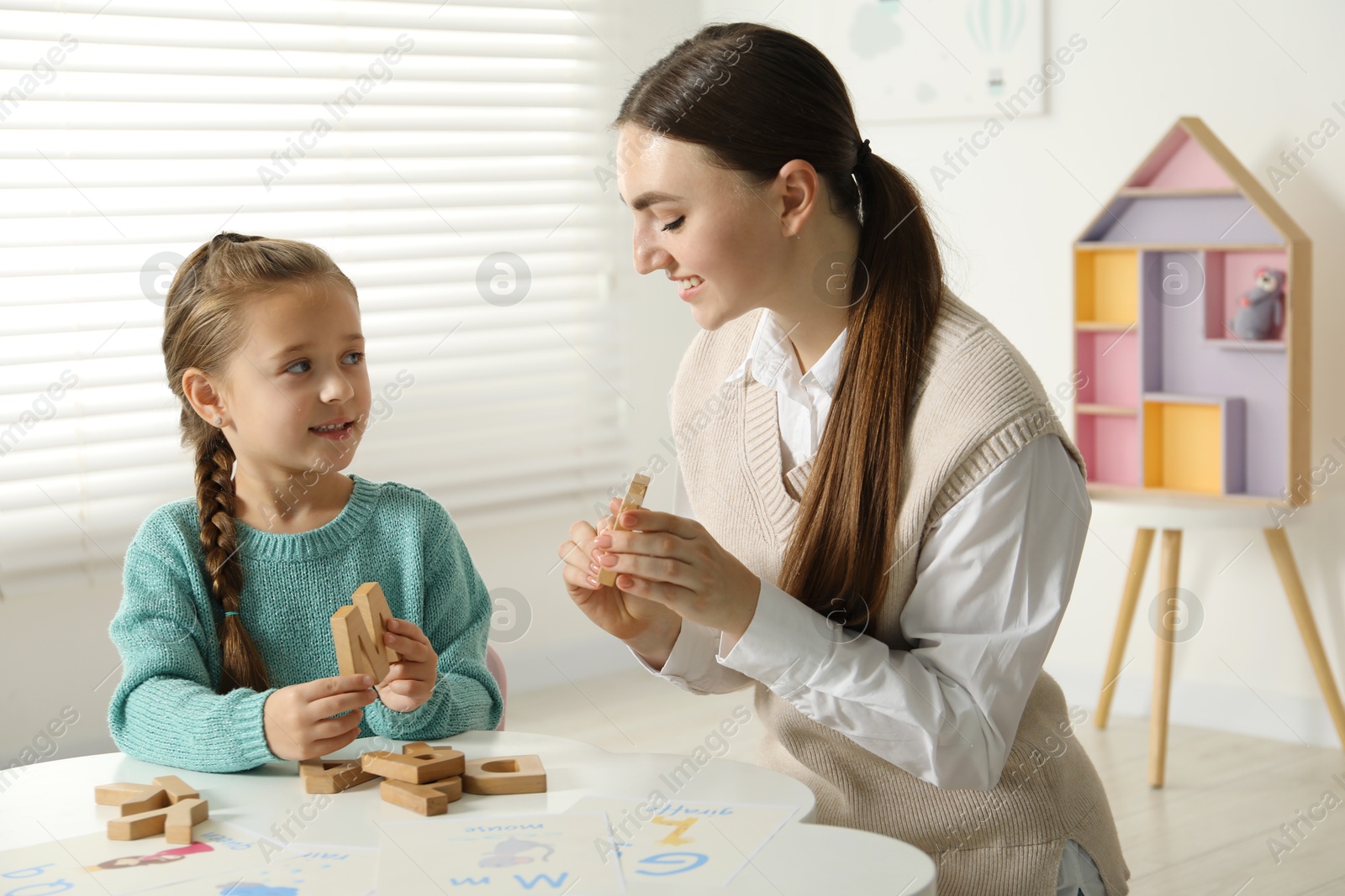 Photo of Speech therapist teaching little girl alphabet with wooden letters at white table indoors