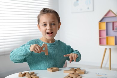 Photo of Learning alphabet. Little girl with wooden letter A at white table indoors