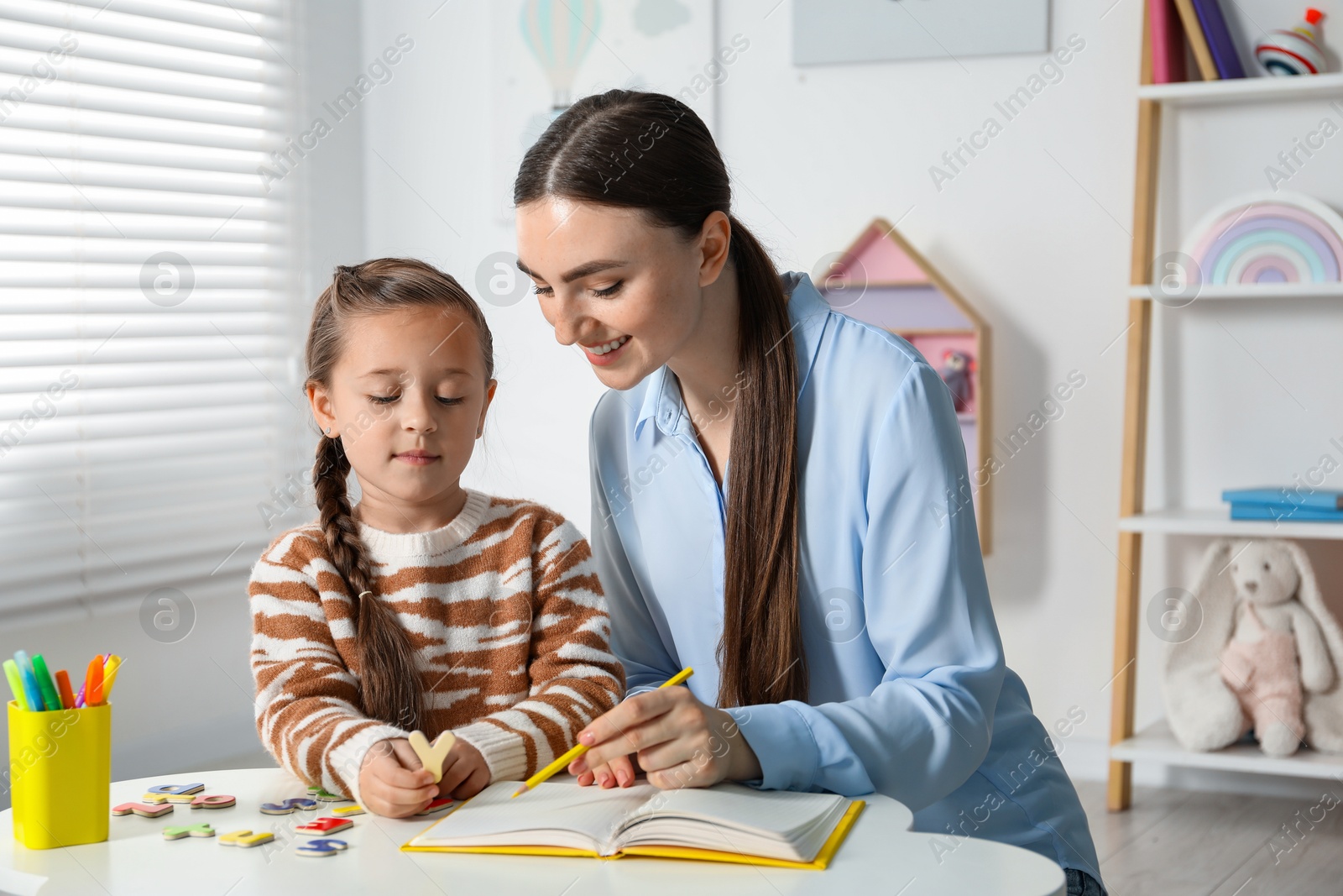 Photo of Speech therapist teaching little girl alphabet with magnetic letters at white table indoors