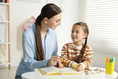 Photo of Speech therapist teaching little girl alphabet with magnetic letters at white table indoors