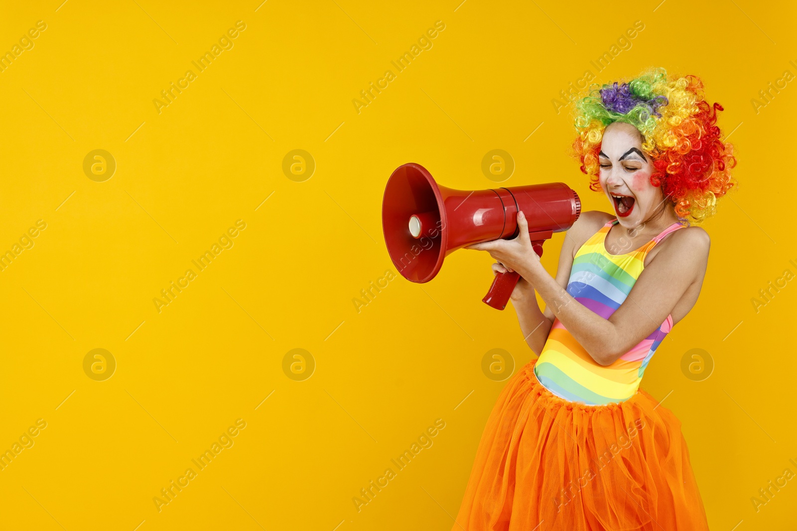 Photo of Cute girl dressed like clown shouting in megaphone on yellow background, space for text. Surprise party