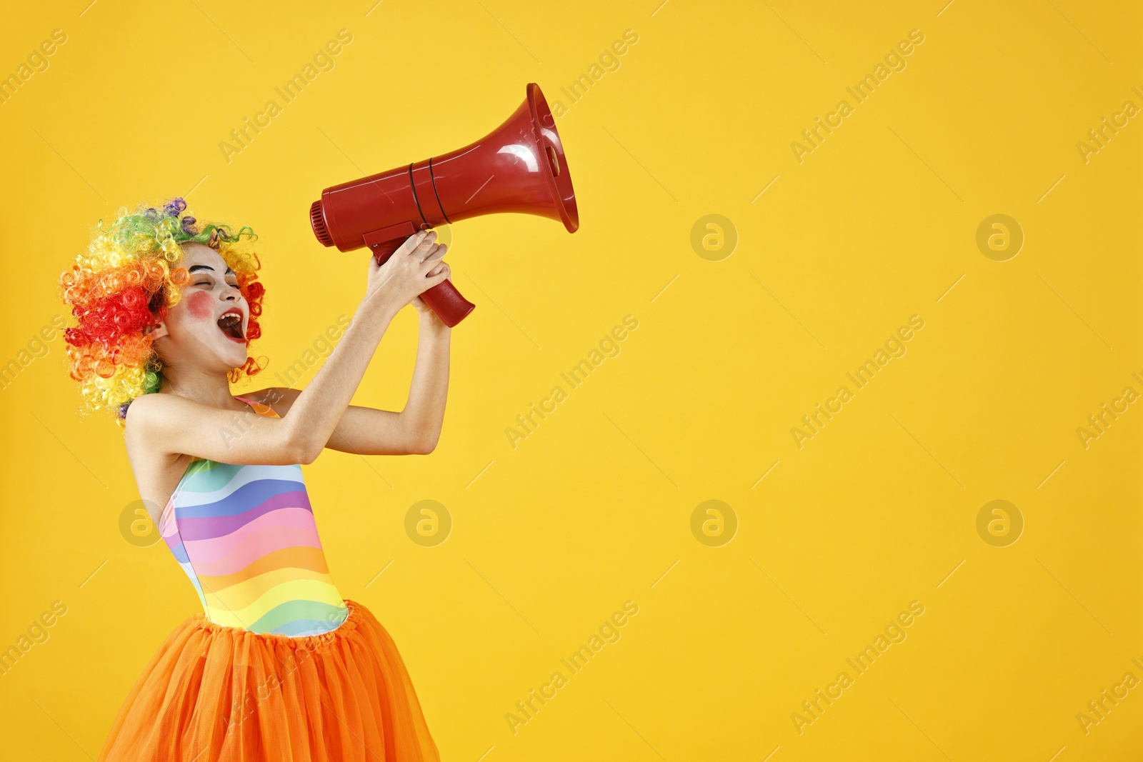 Photo of Cute girl dressed like clown shouting in megaphone on yellow background, space for text. Surprise party