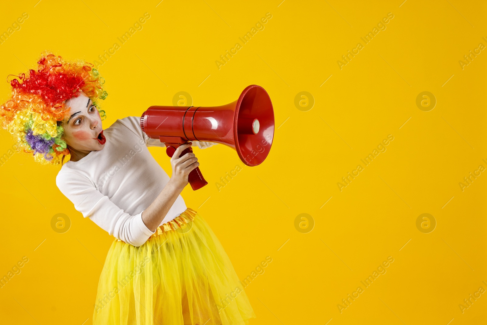 Photo of Cute girl dressed like clown shouting in megaphone on yellow background, space for text. Surprise party