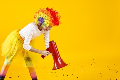 Girl dressed like clown with megaphone and confetti on yellow background, space for text. Surprise party