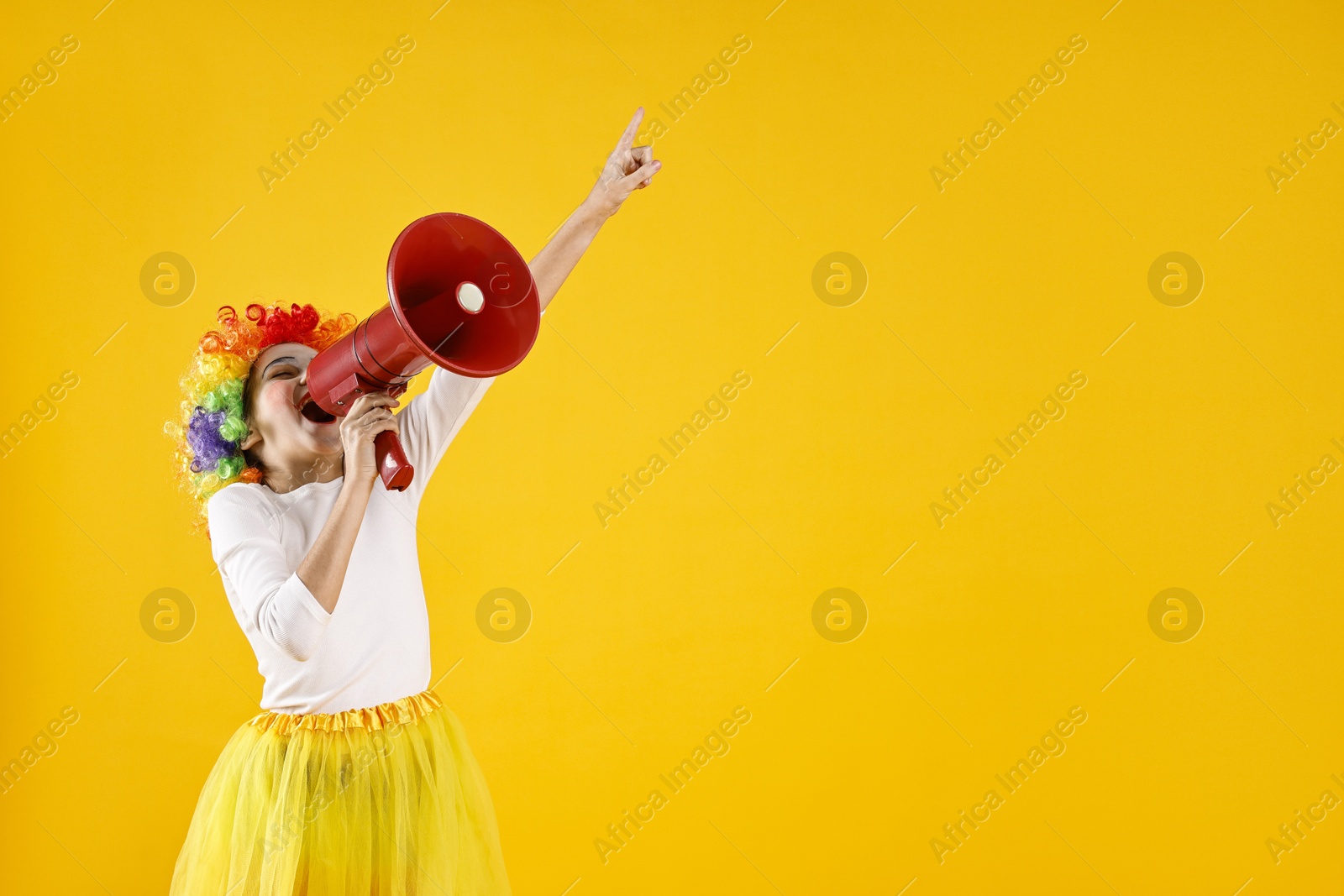 Photo of Cute girl dressed like clown shouting in megaphone and pointing upwards on yellow background, space for text. Surprise party