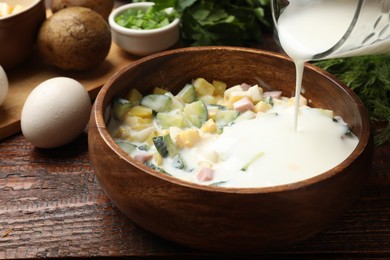 Photo of Making okroshka soup. Adding kefir to bowl with ingredients at wooden table, closeup