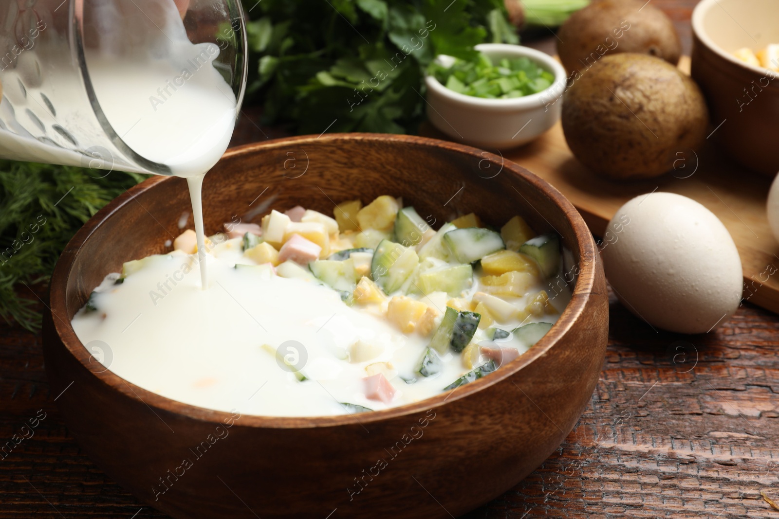 Photo of Making okroshka soup. Adding kefir to bowl with ingredients at wooden table, closeup