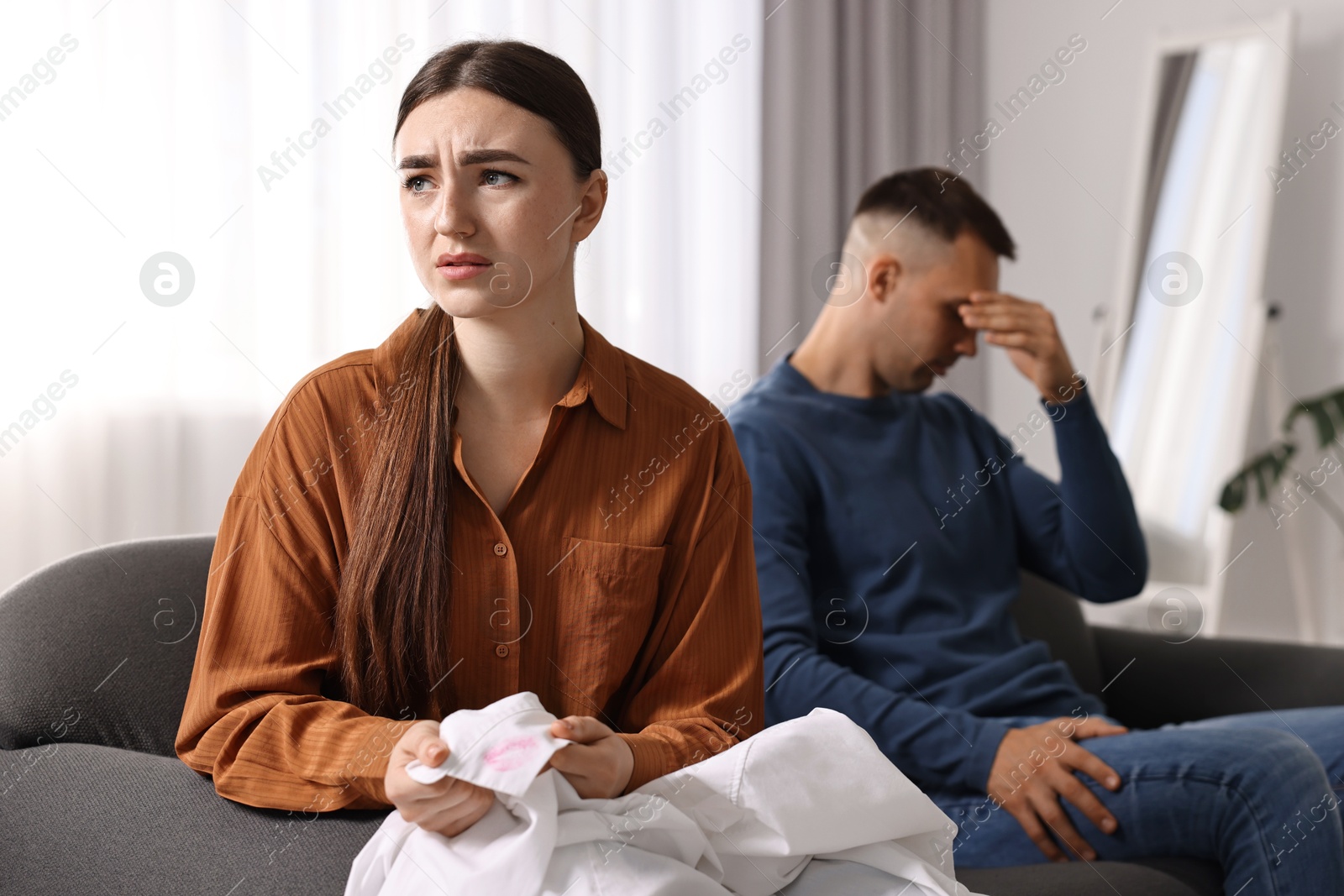 Photo of Frustrated wife holding shirt with lipstick marks as proof of her unfaithful husband at home, selective focus