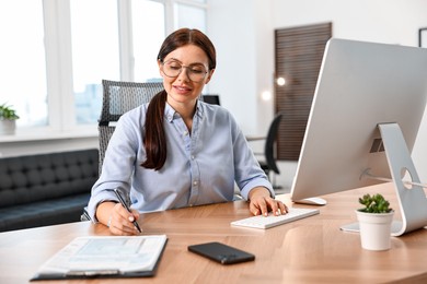 Photo of Woman taking notes while working on computer at table in office