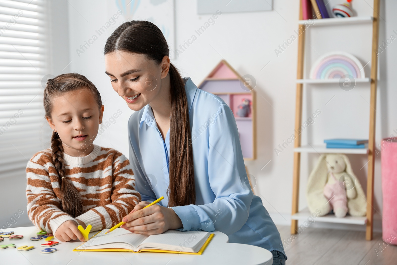 Photo of Speech therapist teaching little girl alphabet with magnetic letters at white table indoors