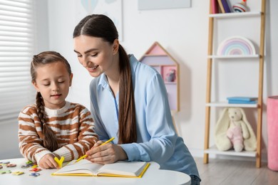 Photo of Speech therapist teaching little girl alphabet with magnetic letters at white table indoors