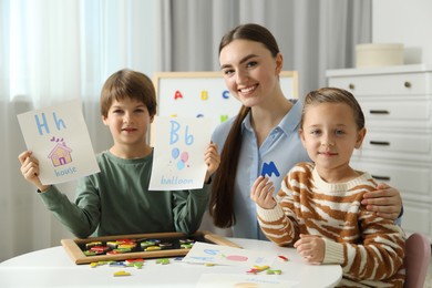 Photo of Speech therapist teaching little kids alphabet with magnetic letters and pictures at white table indoors