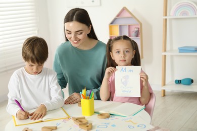 Photo of Speech therapist teaching little kids alphabet at white table indoors