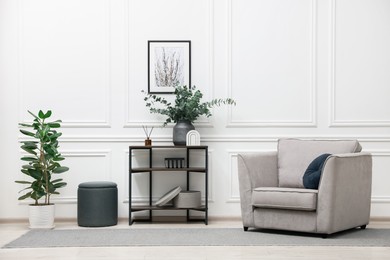 Photo of Console table with decor, houseplant, pouf and armchair near white wall in room. Interior design