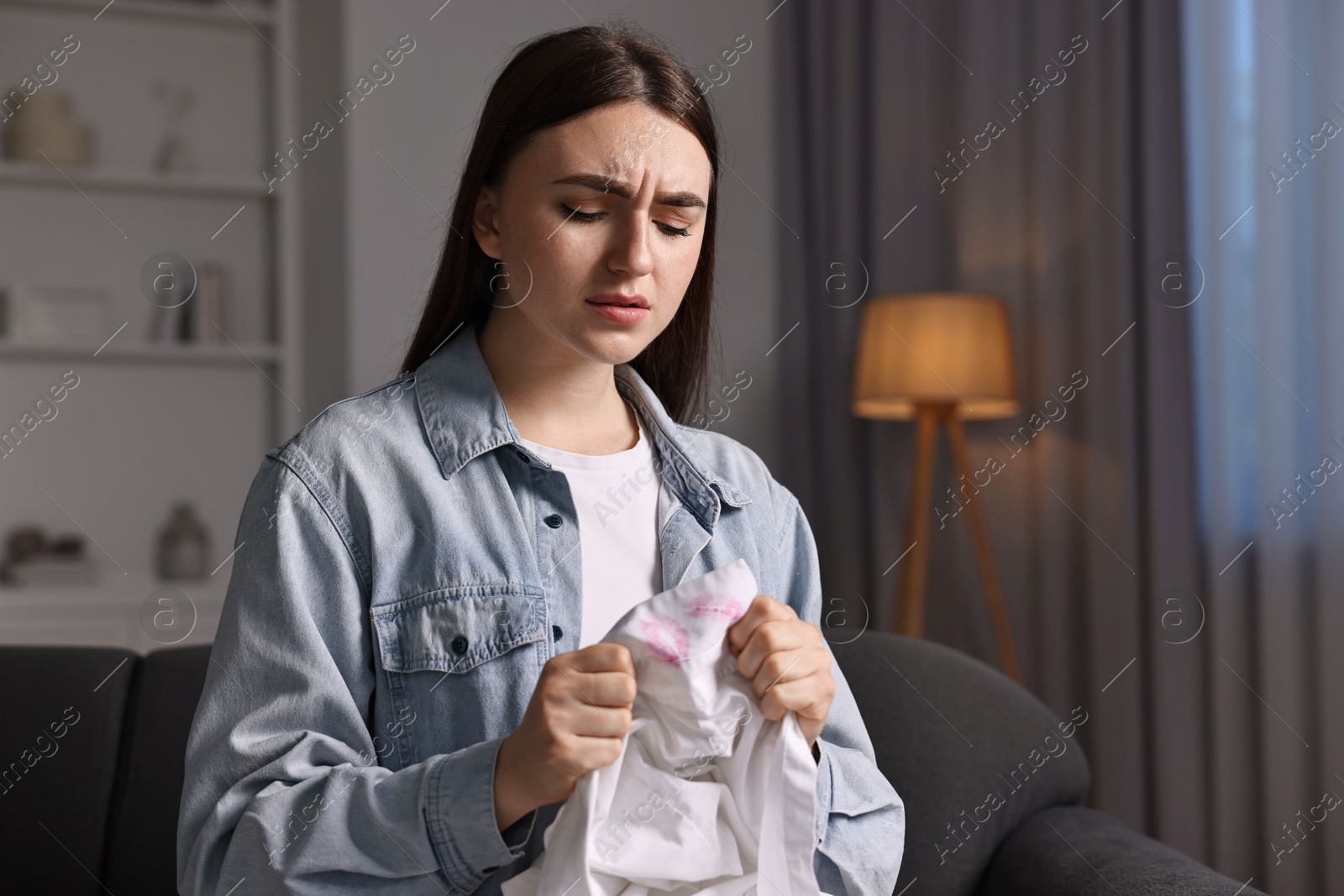 Photo of Frustrated wife holding shirt with lipstick marks as proof of her unfaithful husband at home