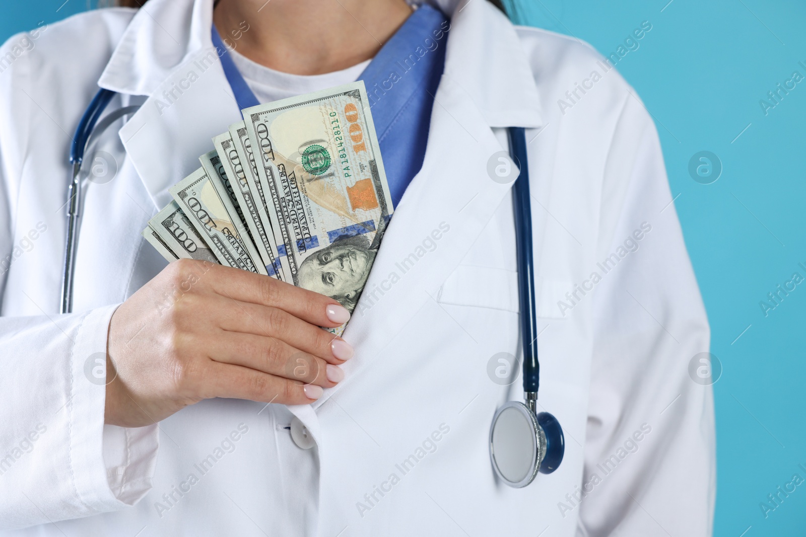 Photo of Corruption concept. Doctor putting dollar banknotes into her coat on light blue background, closeup