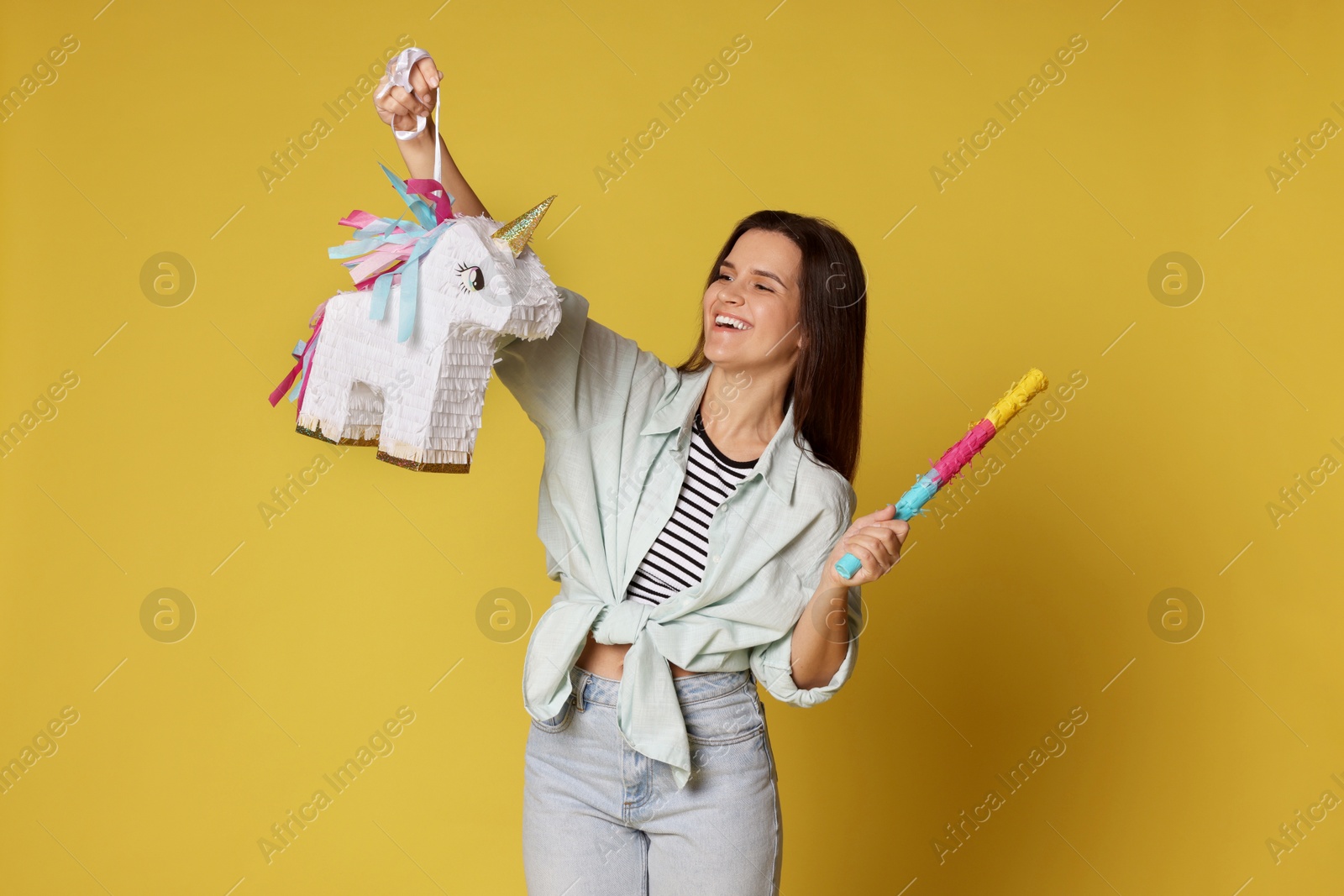 Photo of Happy woman with unicorn shaped pinata and stick on yellow background