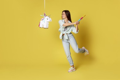 Photo of Happy woman hitting unicorn shaped pinata with stick on yellow background